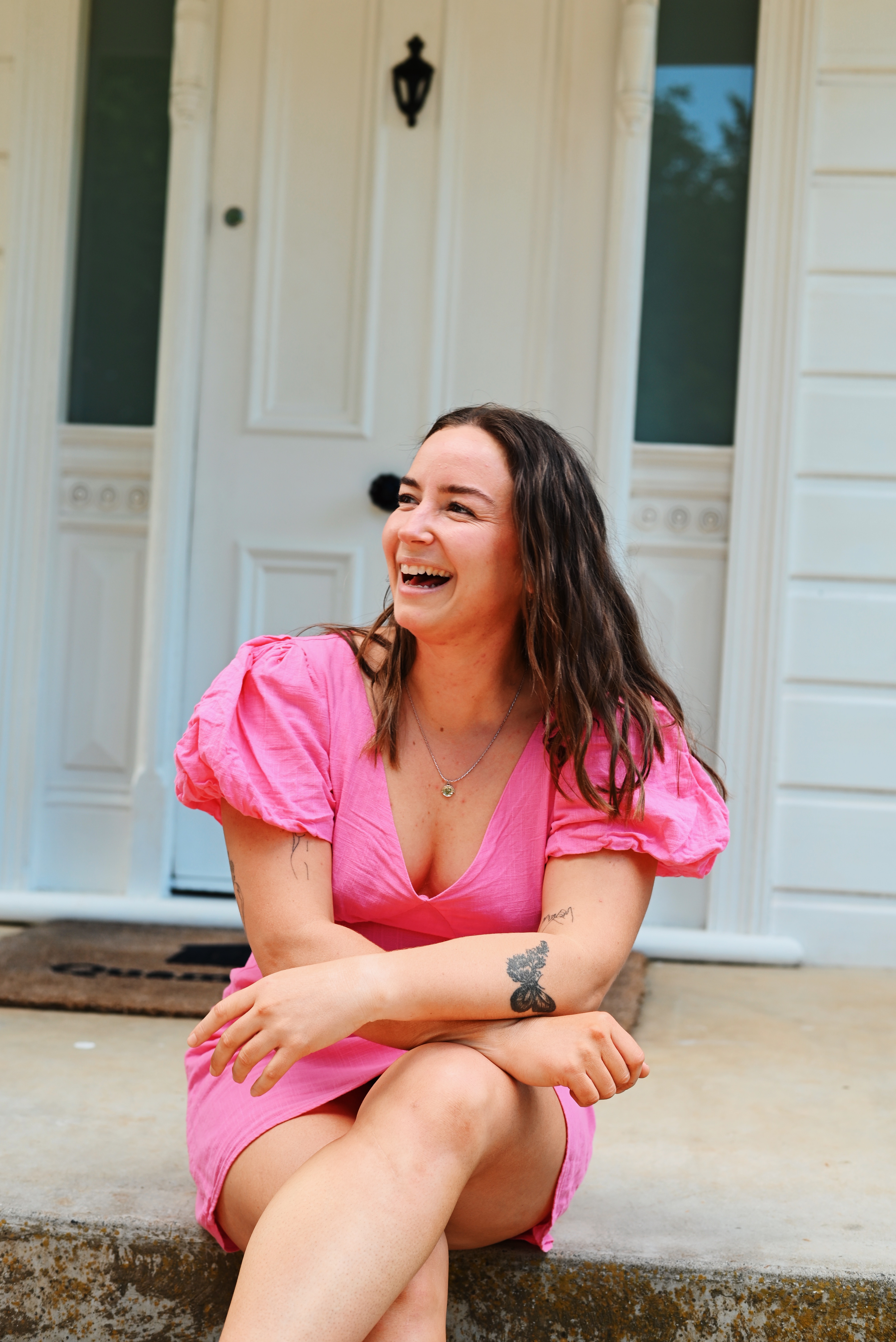 A woman sitting on a doorstep, laughing and wearing a pink dress with puff sleeves, in front of a white door.
